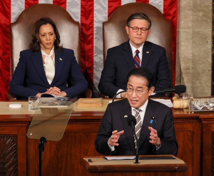 Prime Minister Kishida stands at a lecturn giving a speech, Vice President Harris and Speaker Johnson are seated behind him.