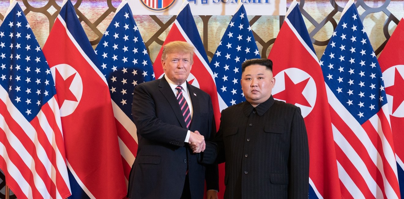 Donald J. Trump stands next to Kim Jong Un as they shake hands and pose in front of U.S. and North Korean flags