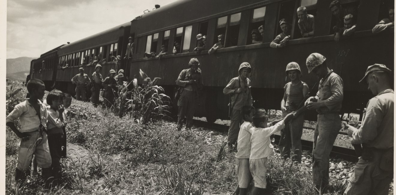 A black and white photo of U.S. Soldiers standing next to a stopped train, handing out candy to South Korean children.