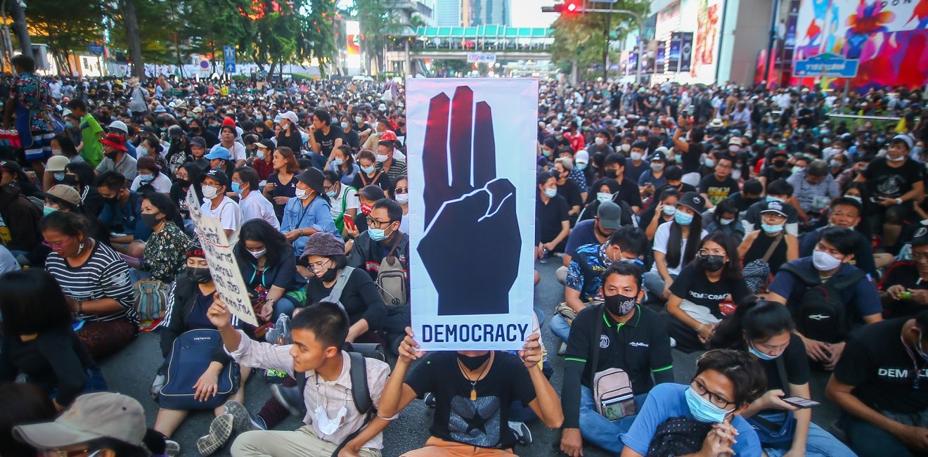 A crowd of demonstrators sit in a roadway, with one in the center holding a sign that reads "democracy"