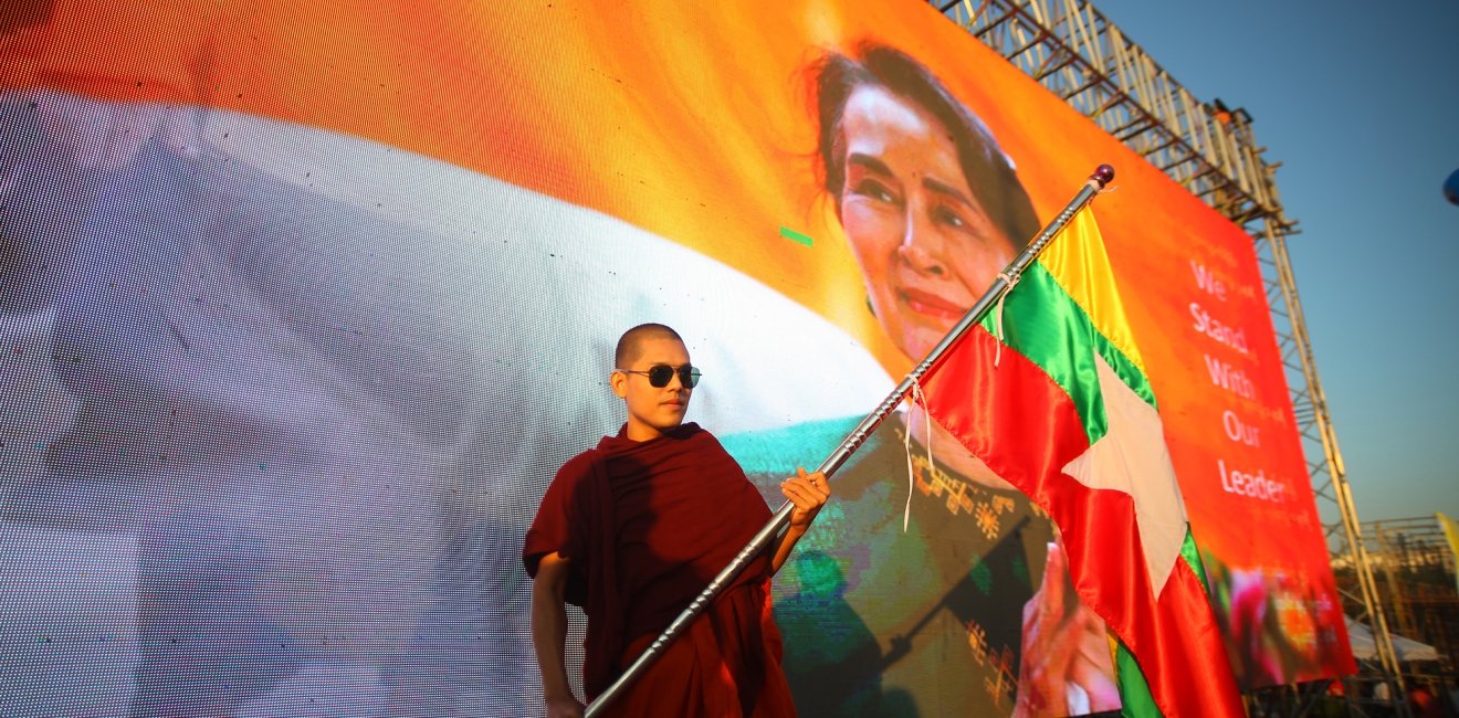 A man in red robes stands with a flag in front of a banner of Aung San Suu Kyi