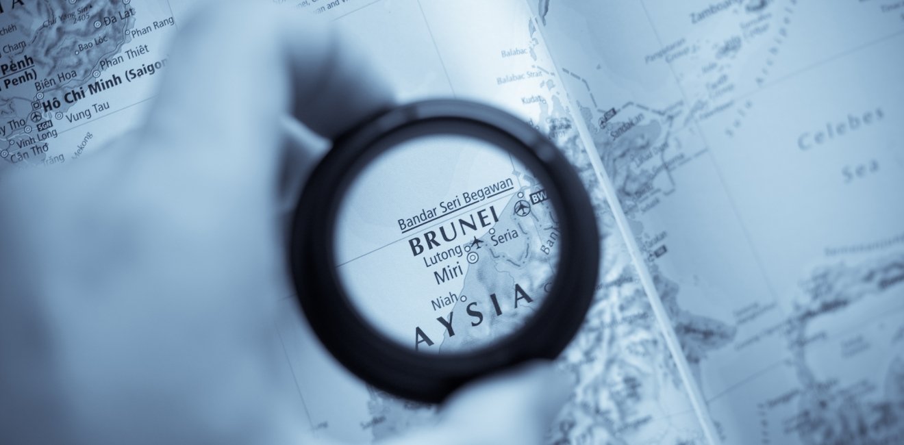 A hand holds a magnifying glass that is circling Brunei on a map.