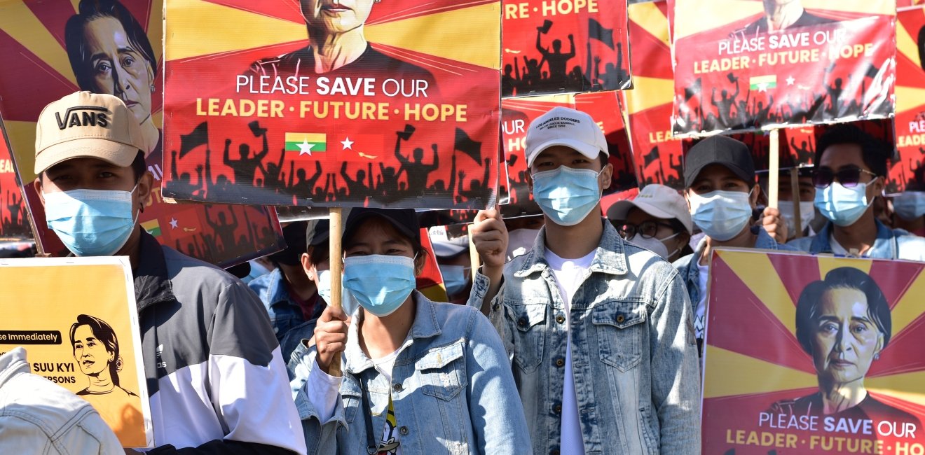 A crowd of protestors wearing surgical masks carry signs reading Please Save our Leader - Future - Hope with a photo of Aung San Suu Kyi