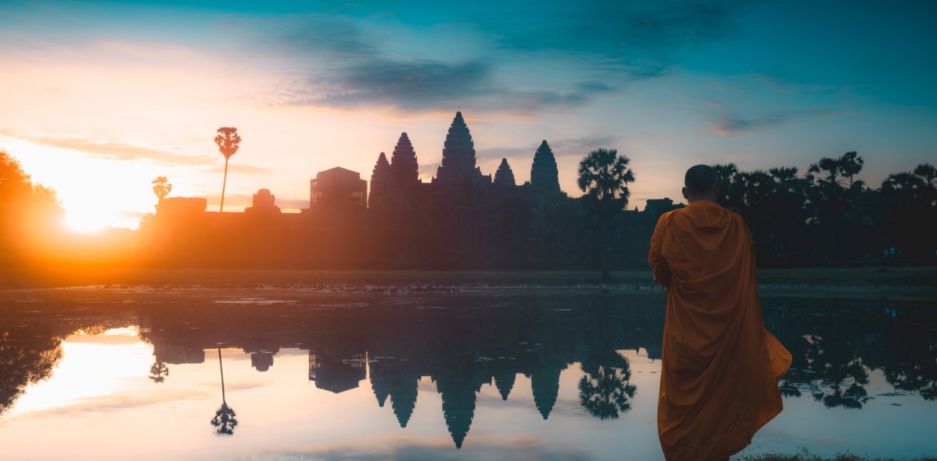 A monk stands in the foreground looking out at a temple at sunrise.