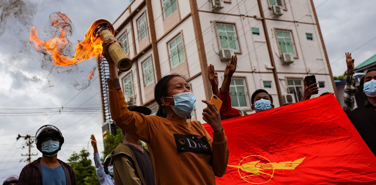 A group of protestors march with the flag of the National League for Democracy, one of them is carrying a lit torch.