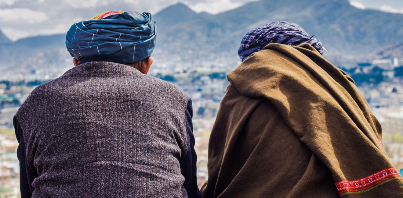 Two men sit with their backs to the camera, looking out at the Afghan mountains.