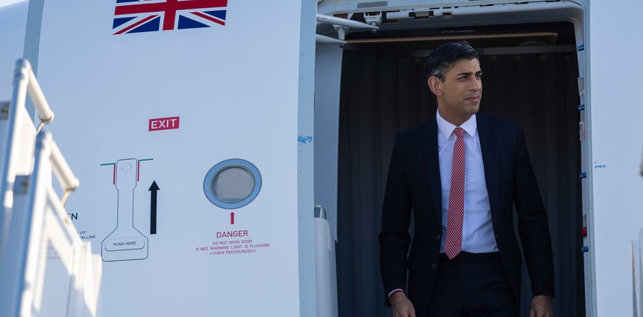 Prime Minister Rishi Sunak exits a plane with a UK flag visible on the door.