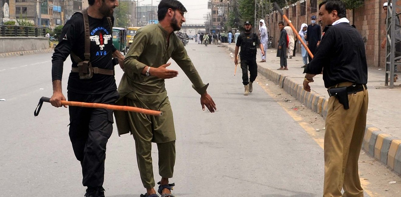 Two men with weapons are detaining another man on the street as people look on.