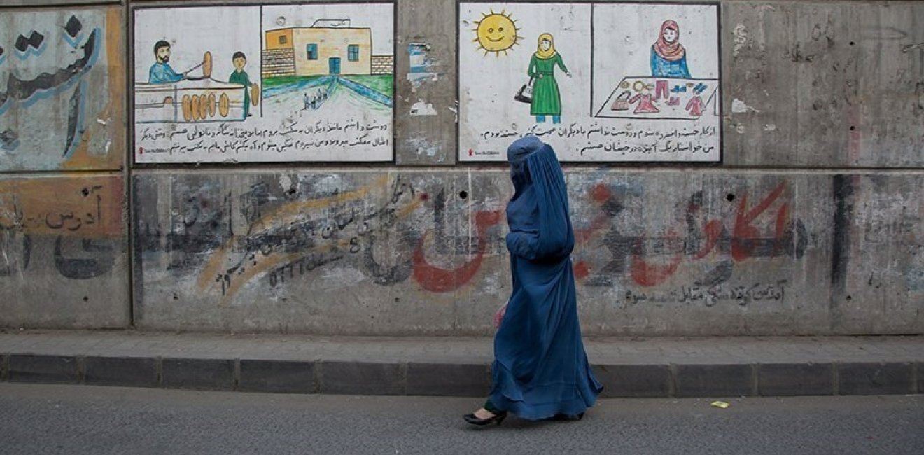 A woman in a blue burqa walks along an empty street in front of murals and graffiti.