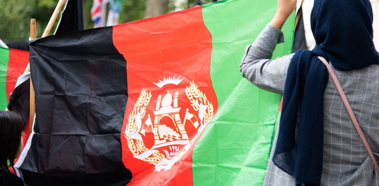 Two people with their backs to the camera are holding a large Afghan flag.