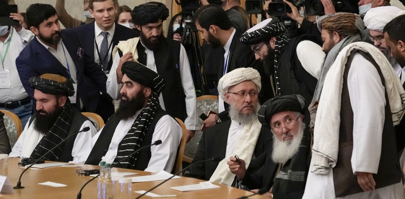 A group of Taliban officials sitting at a conference table with press behind them.