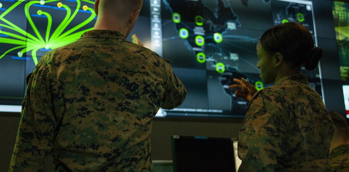 Two soldiers have their back to the camera while looking at a green and blue display of digital information.