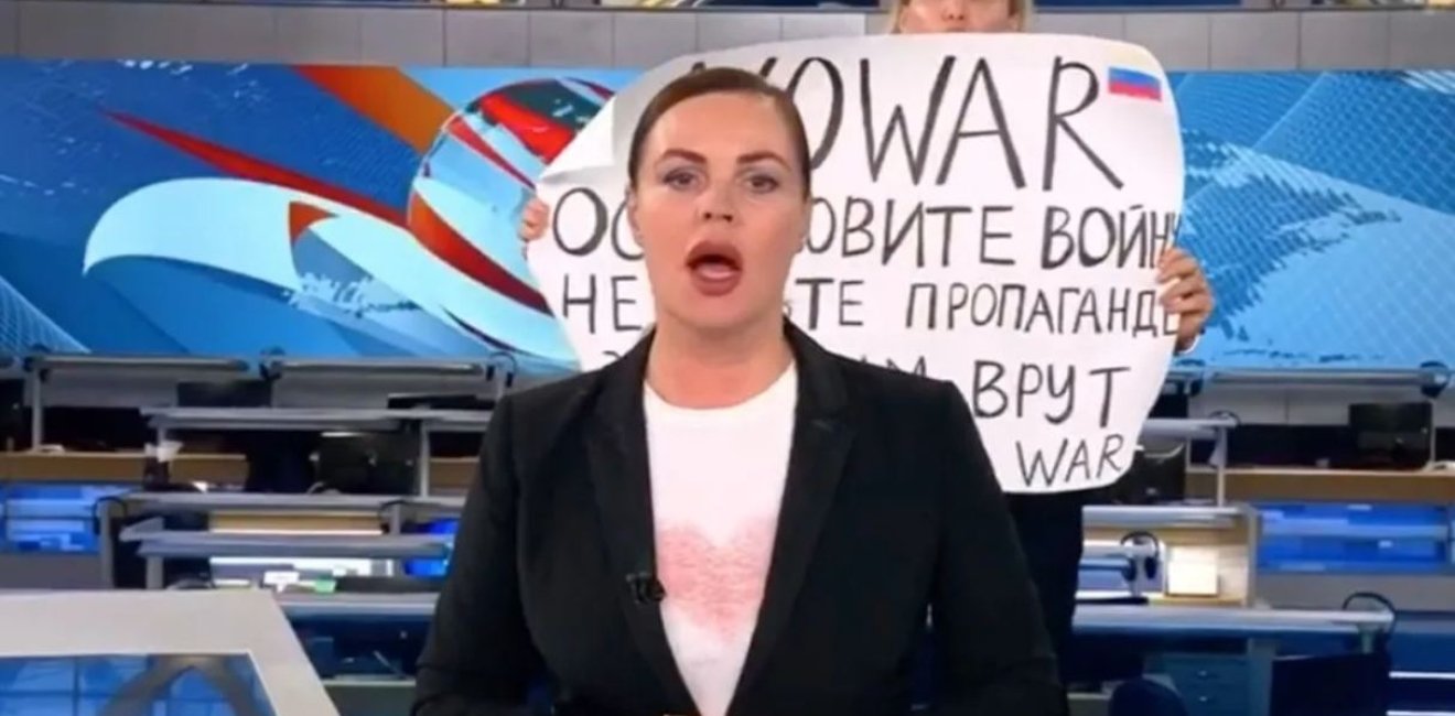 A protester holding sign behind a newscaster 