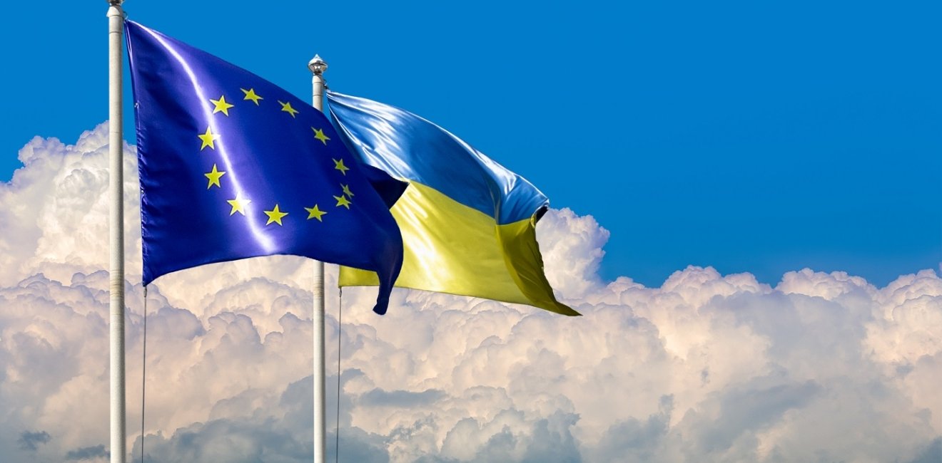 Two flags of Ukraine and the European Union on flagpoles at blue cloudy sky.