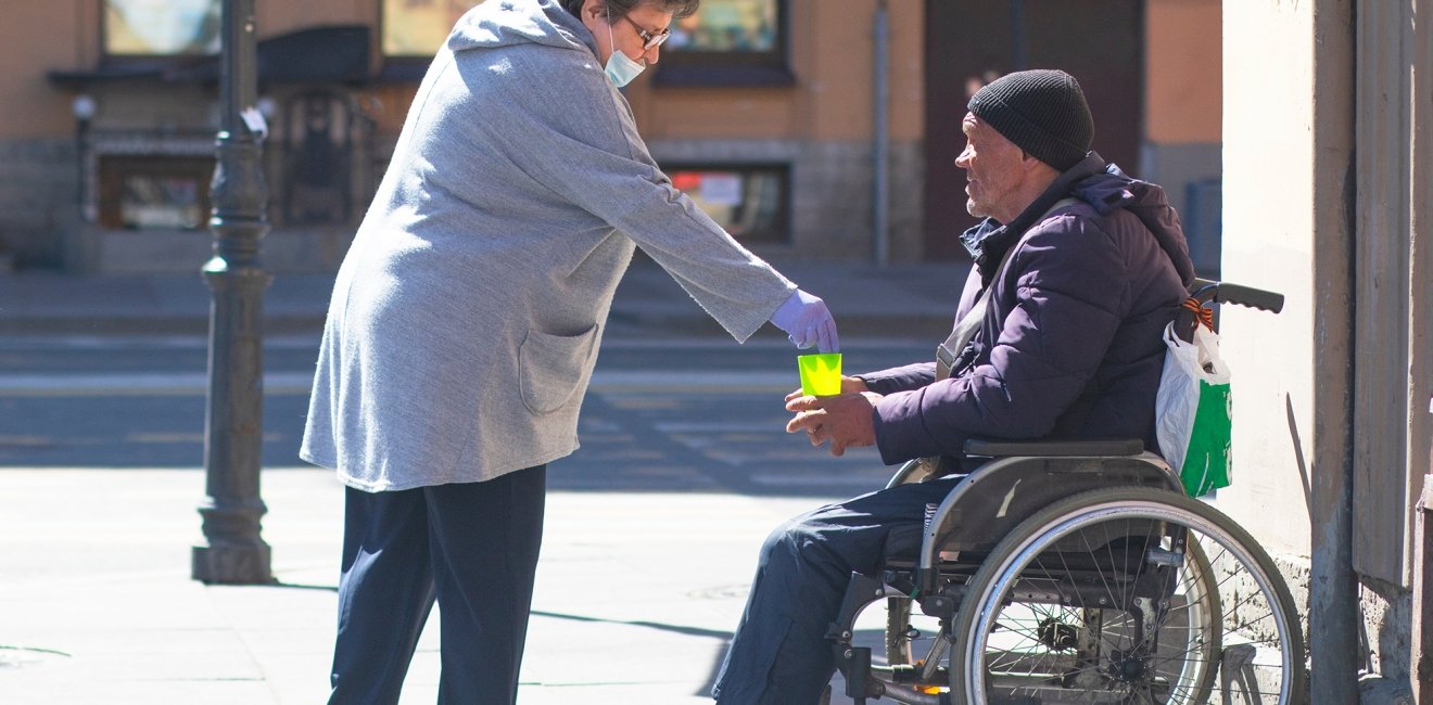 Adult elderly woman in a protective medical mask and gloves gives money to a man in a wheelchair in St. Petersburg.