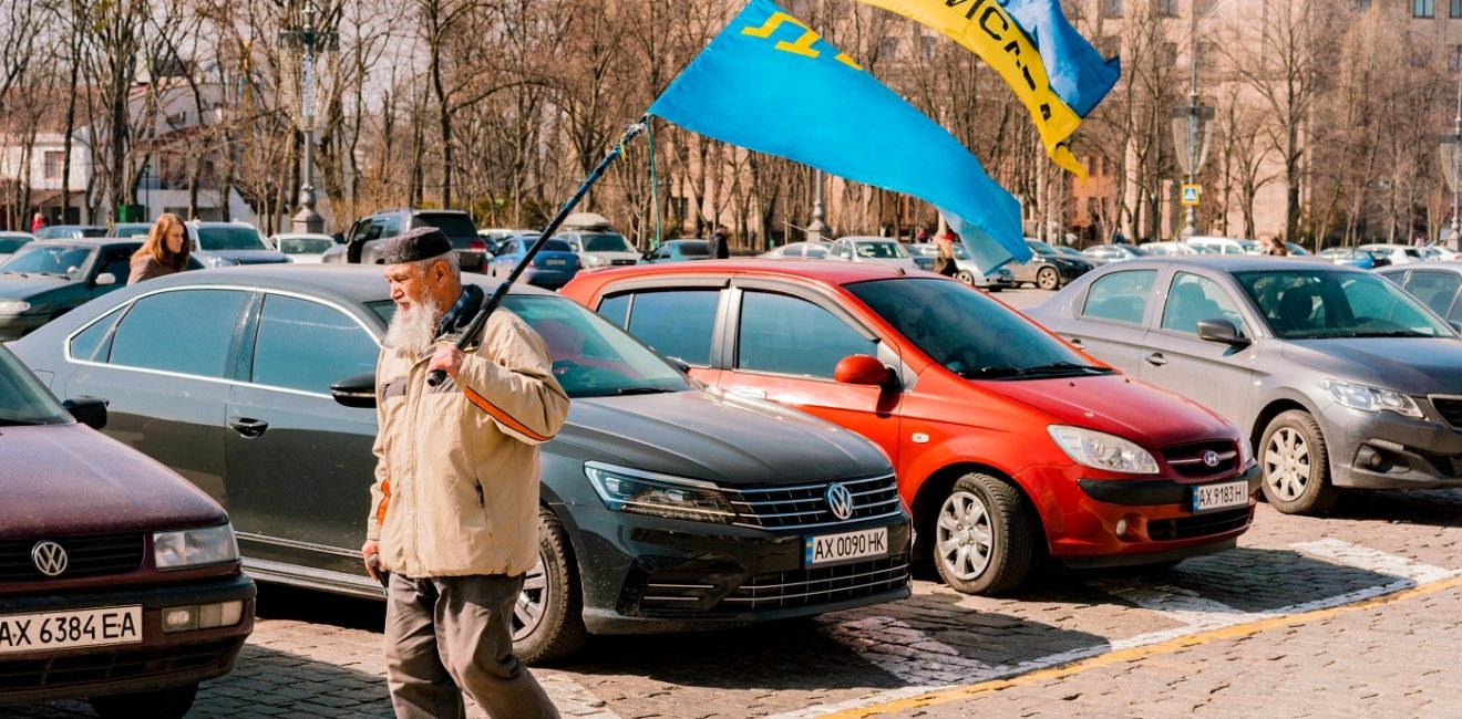 Crimean Tatar man with big beard and traditional cap walks on the city's street with Crimea Tatars and Ukrainian flags. 