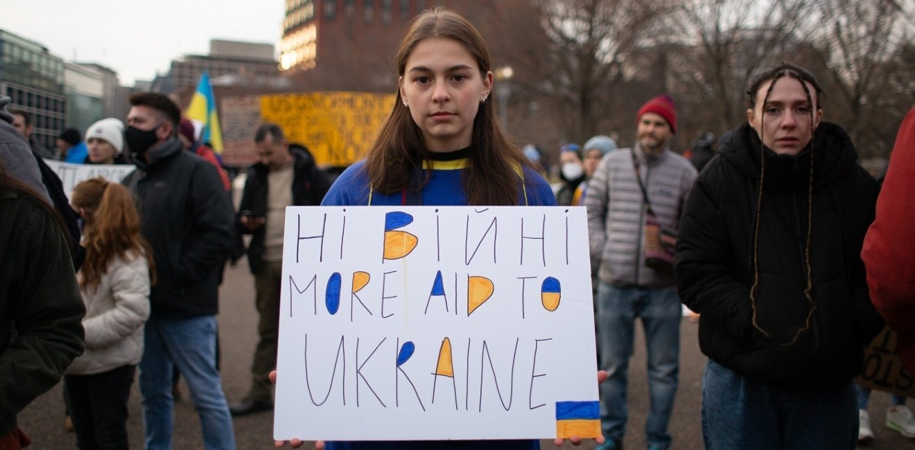 Young Person holding sign with text "Ні війні More Aid To Ukraine"