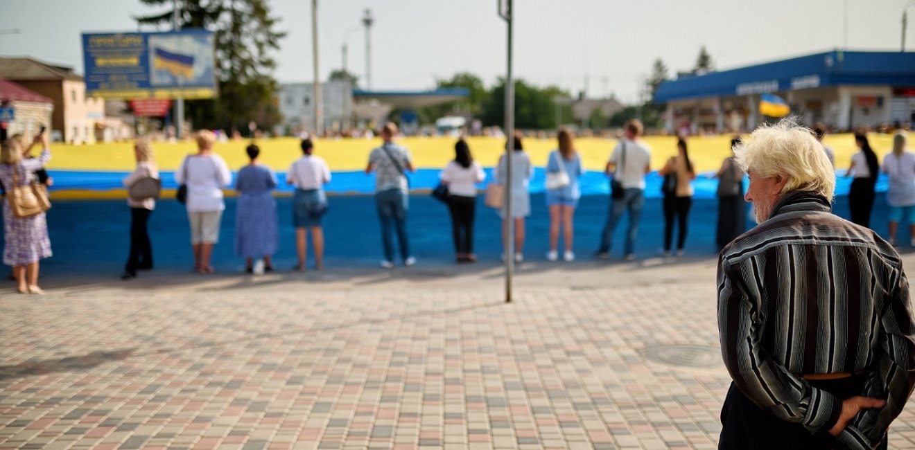 An older man observes a group of people holding a large Ukrainian Flag