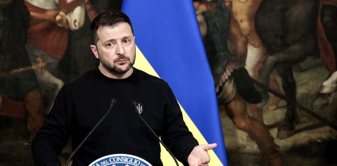 Volodymyr Zelensky standing in front of a Ukrainian flag and a painting of ancient warriors