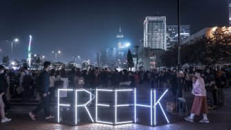 A group of protestors are gathered at night, lights spell out Free HK in front of them.