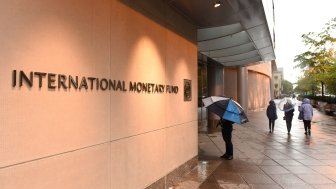 People with umbrellas walking past a building with a sign that says International Monetary Fund.