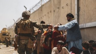 A man in a military uniform hands a bottle of water to an Afghan civilian waiting in a crowd of people.