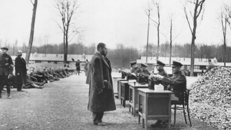 Gestapo officials recording data on incoming prisoners at a German concentration camp. Many others are seated on the ground