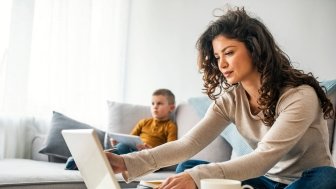 Smiling mom working at home with her child on the sofa while writing an email. 