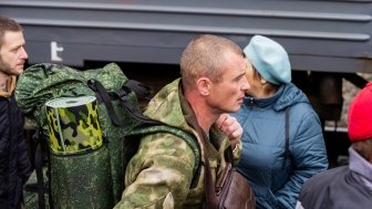 A white man in green camoflage, wearing a green camoflage backpack, walks in front of a train. 