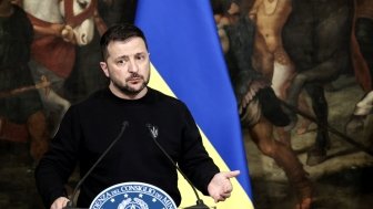 Volodymyr Zelensky standing in front of a Ukrainian flag and a painting of ancient warriors