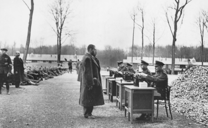 Gestapo officials recording data on incoming prisoners at a German concentration camp. Many others are seated on the ground