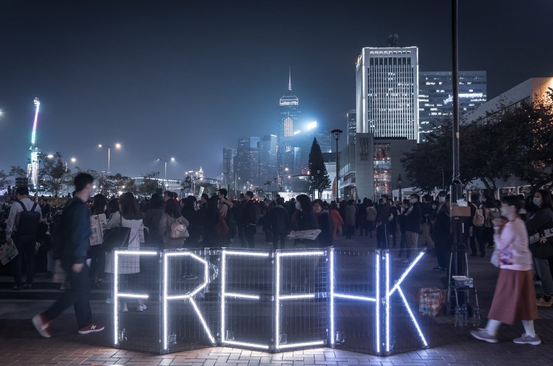 A group of protestors are gathered at night, lights spell out Free HK in front of them.