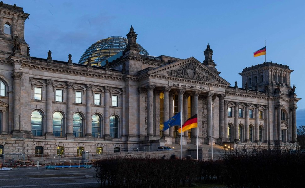 The Bundestag building at dusk, with German and EU flags waving in front. 