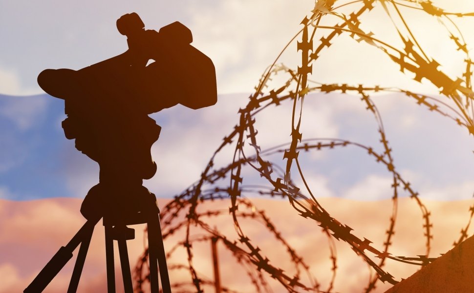 A composite photo with the Russian flag in the background, and barbed wire and a television news camera in the foreground