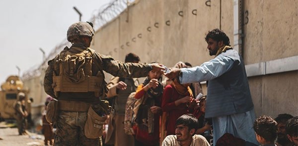 A man in a military uniform hands a bottle of water to an Afghan civilian waiting in a crowd of people.