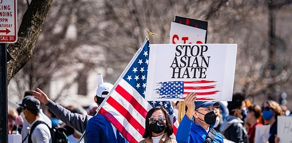 A man stands in a group of protestors wearing a masks, holding a sign with an American flag on it that says Stop Asian Hate.