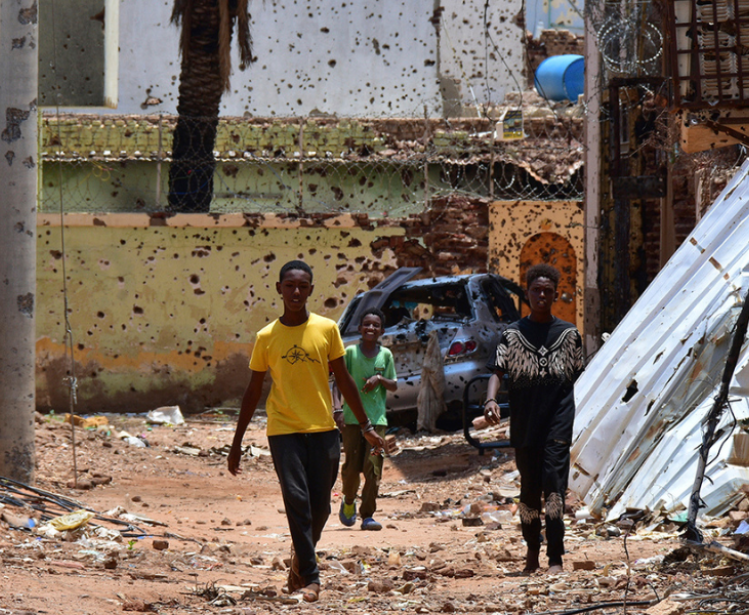 Young people walk along a street marked by destruction. A bloody power struggle has been raging in Sudan for more than 16 months, triggering a refugee crisis. Mudathir Hameed/picture-alliance/dpa/AP Images.